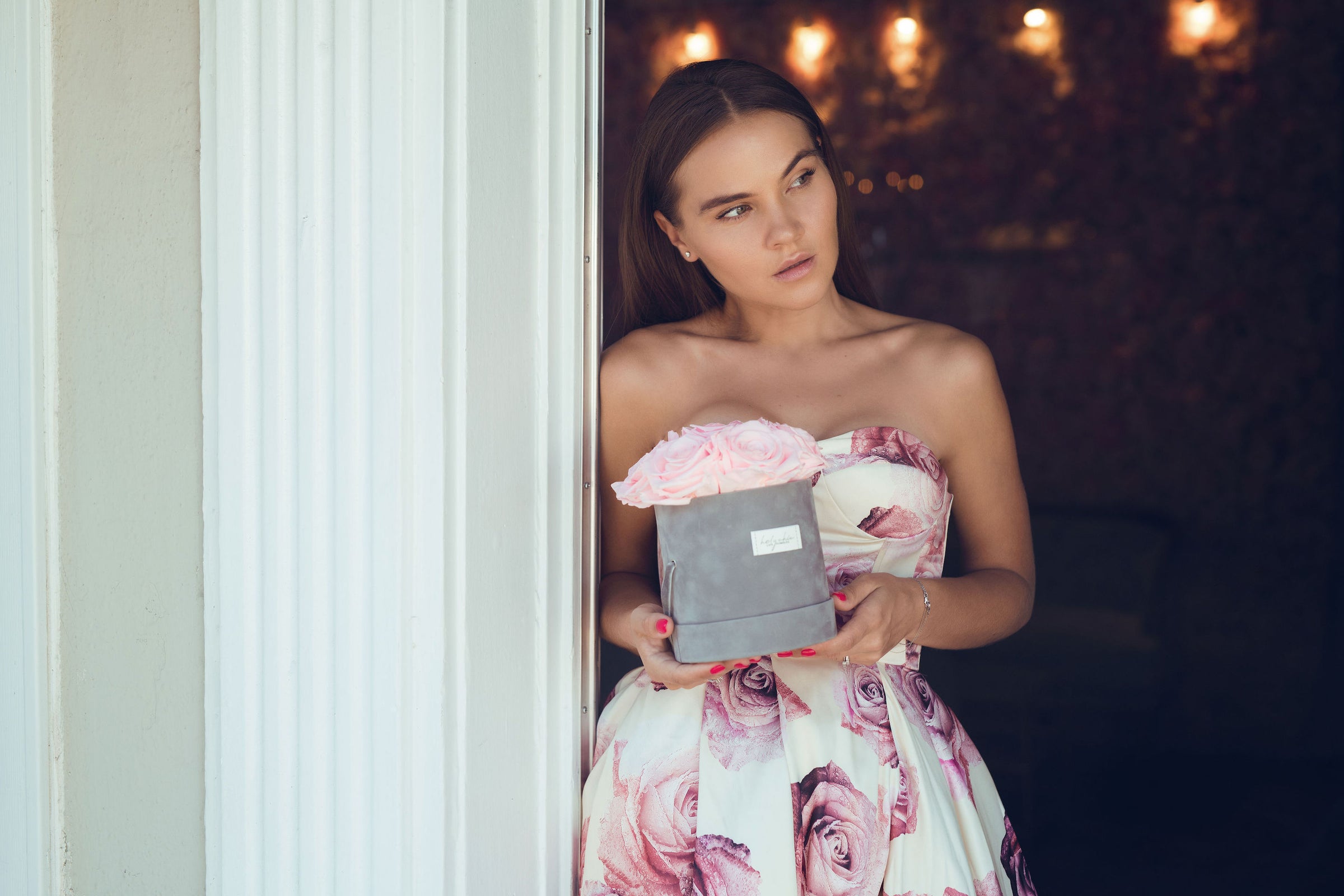 Girl in a flower print white dress holding a grey velvet box of pink roses in her hand